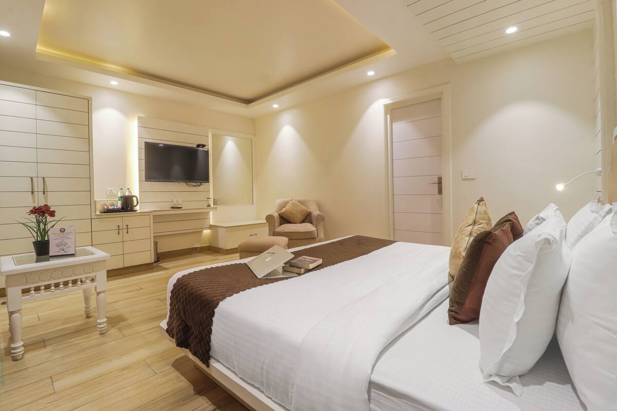 A modern hotel room with a large bed, a TV mounted on the wall, a bedside table with a lamp, and a chair.