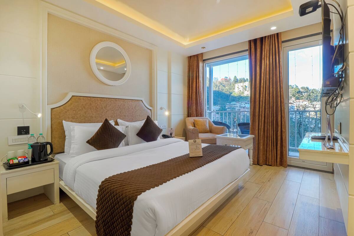 A hotel room with a large bed, a bedside table with a kettle, a TV mounted on the wall, and a view of the city through large windows.