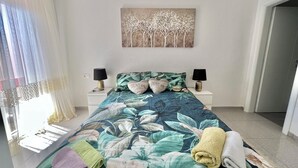 Een slaapkamer met een bloemenprint bedsprei, twee bedlampen, een schilderij aan de muur en uitzicht buiten door dunne gordijnen.