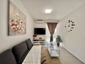 A modern living room with a sofa, a flat-screen TV, a decorative painting, a clock, and a potted plant. - Apartman Ana 1 (Dramalj)