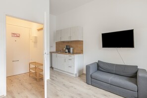 Una cocina moderna con armarios blancos, una encimera de madera y una cafetera. Un sofá gris se encuentra frente a un televisor montado en la pared.