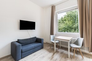 Ein Zimmer mit einem an der Wand befestigten Flachbildfernseher, einem grauen Sofa, einem kleinen Holztisch mit zwei Stühlen und einem Fenster mit Vorhängen.