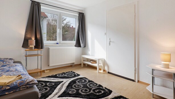 A bedroom with a bed, a window with curtains, a bedside table with a lamp, a white bench, and a radiator. - Apartment 'C' in Miltenberg (Miltenberg)