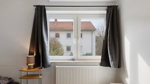A room with a window, a radiator, a lamp, and a view of a house. - Apartment 'C' in Miltenberg (Miltenberg)