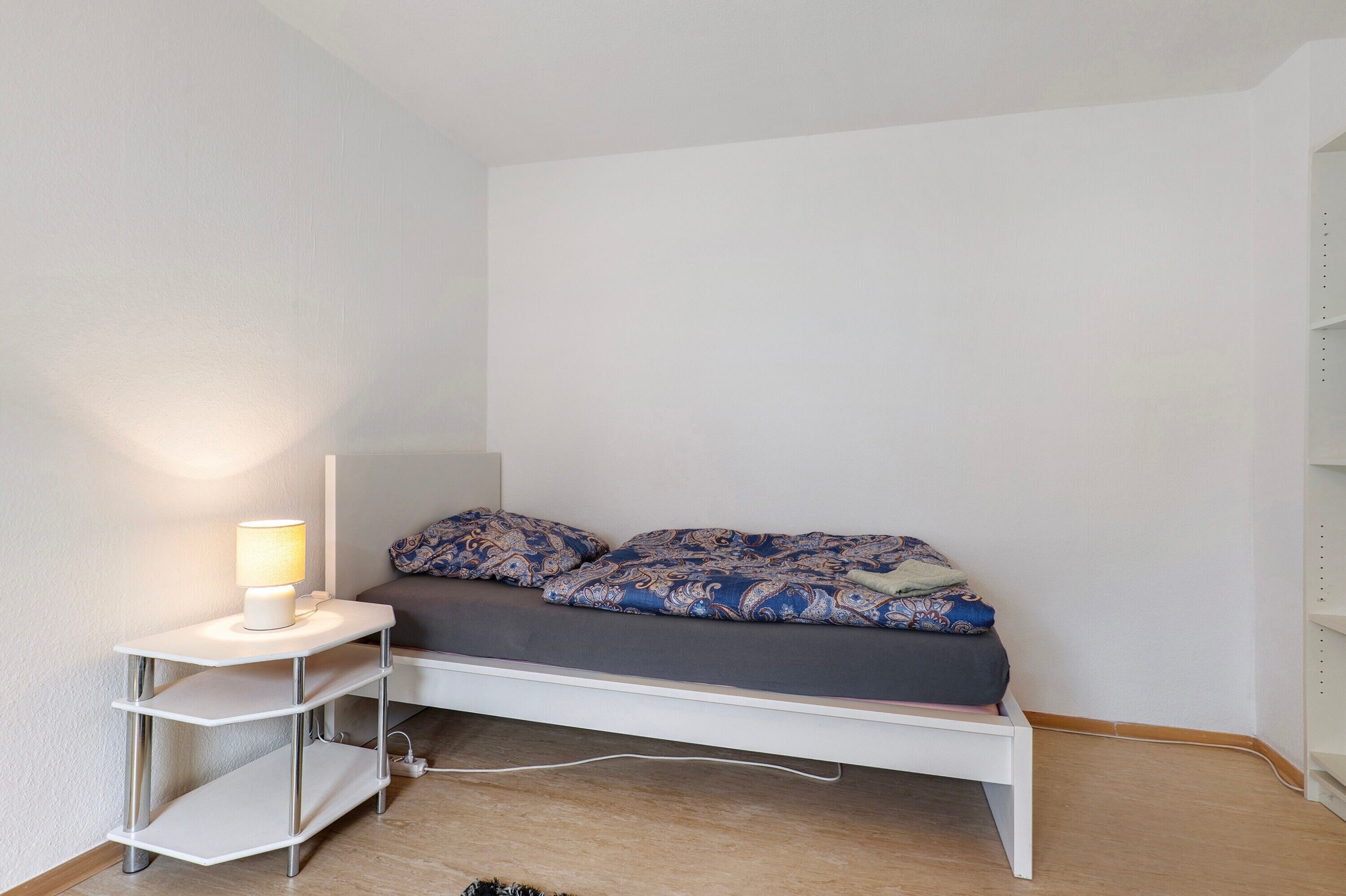 Ein kleines Zimmer mit einem Einzelbett, einem weißen Kopfteil, einem Nachttisch mit Lampe und einer gesteppten Bettdecke.