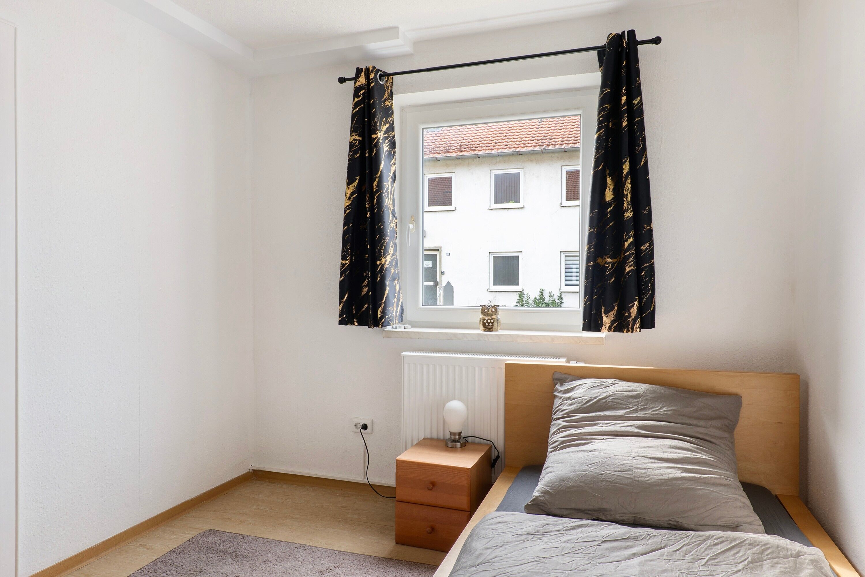 A bedroom with a bed, a nightstand, a lamp, a window with curtains, and a radiator.