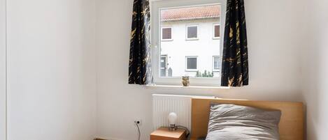 A bedroom with a bed, a nightstand, a lamp, a window with curtains, and a radiator.