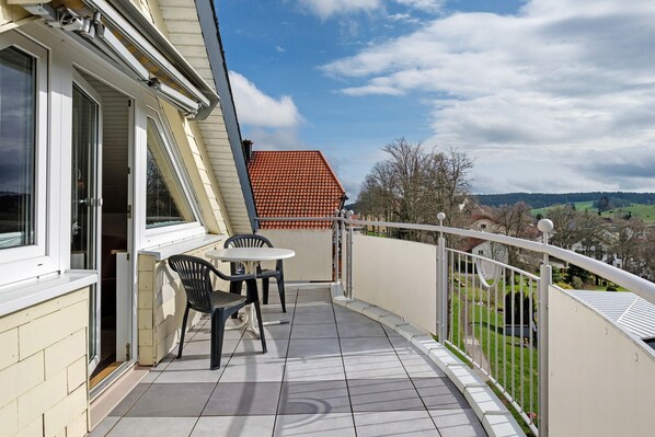 A balcony with a table and chairs, a view of the countryside, and a red-roofed building in the background.