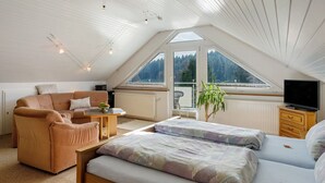 Ein gemütliches Zimmer mit schrägen Wänden, einer Couch, einem Bett, einem Fernseher und einem Fenster mit Blick auf Bäume.