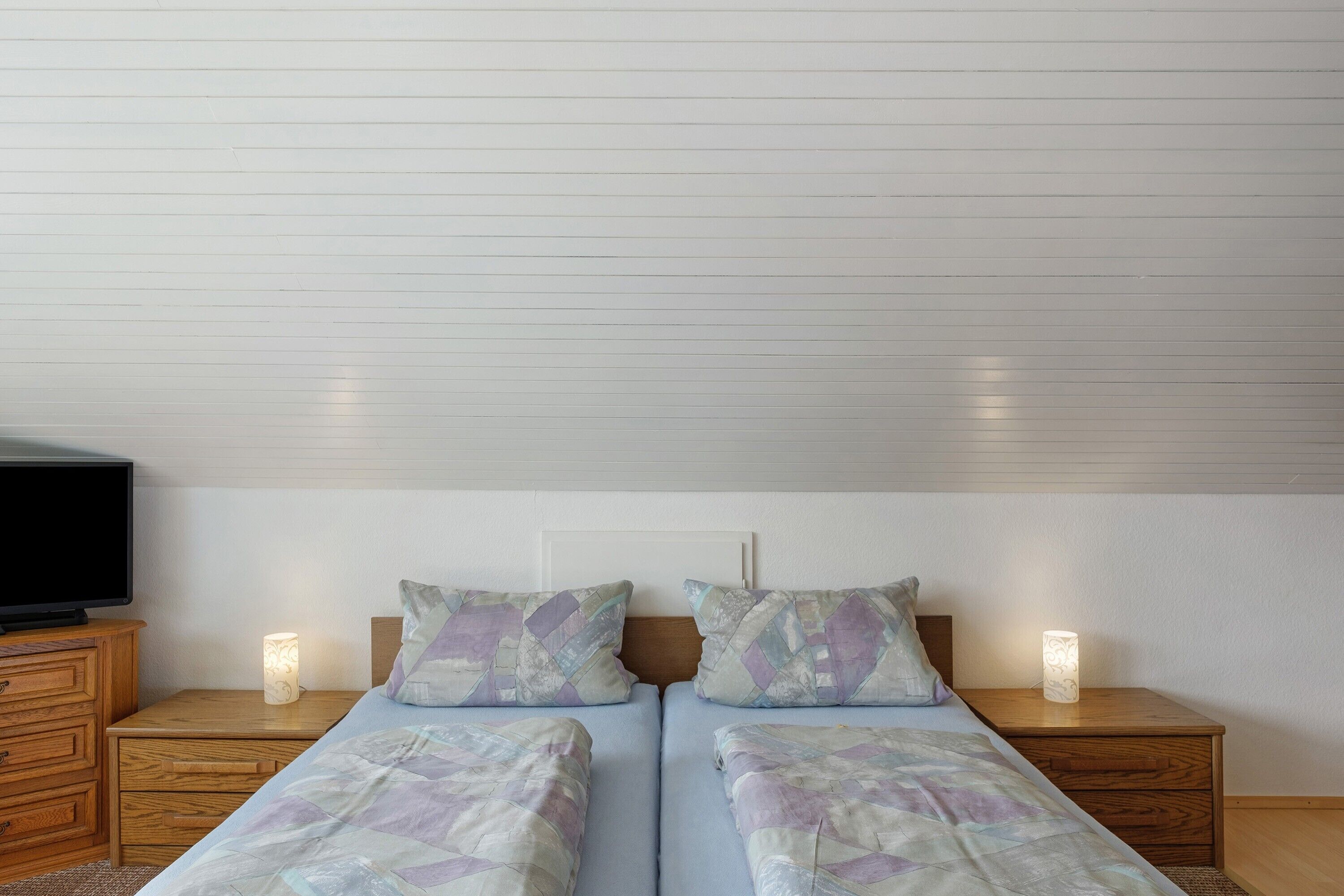 A double bed with patterned bedding, two bedside tables with lamps, a wooden dresser, and a television on the left.
