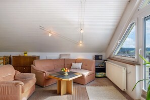 Un salon chaleureux avec un plafond haut, une table en bois sur laquelle sont disposés des fruits, un canapé rose et une vue dégagée sur l’extérieur grâce à une grande fenêtre.