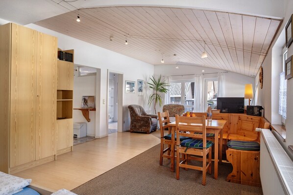 A spacious living area with a wooden ceiling, a dining table with chairs, a sofa, a television, and a window with curtains.