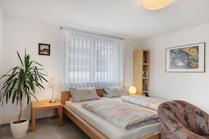 Une chambre à coucher avec un lit, une table de chevet, une lampe, une plante en pot, une chaise, une bibliothèque et un tableau accroché au mur.