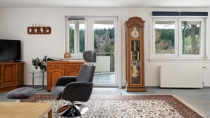 A living room with a television, a large wooden cabinet, a grey armchair, a tall grandfather clock, and a view through the windows. - Studio 'Hörnleberg, 80qm' with Balcony and Wi-Fi (Schönwald im Schwarzwald)