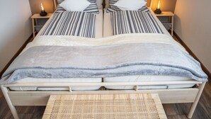 Una cama doble con ropa de cama a rayas, dos mesitas de noche con lámparas, y un banco tejido al pie de la cama.