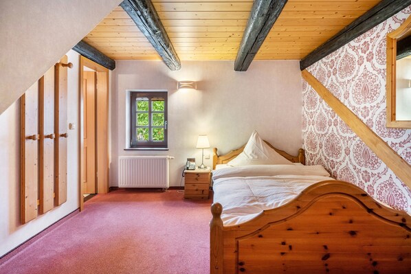 A bedroom with a wooden bed, a nightstand, a lamp, a window with a view of greenery, and a radiator.