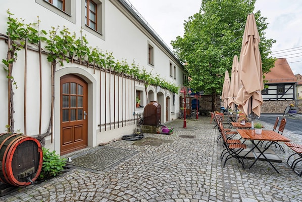 A cobblestone courtyard with outdoor seating, a barrel, and a building with arched windows and a door.