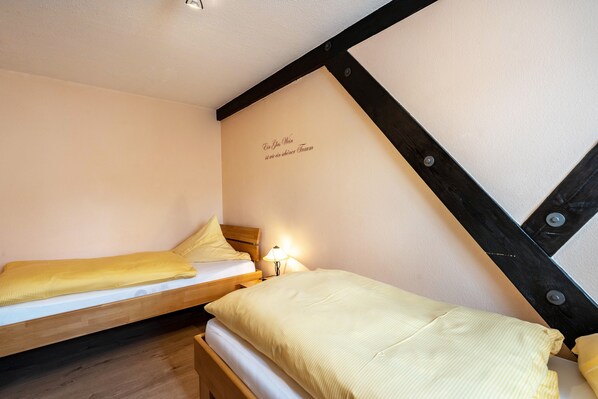 A small, cozy room with two single beds, a nightstand with a lamp, and a wall with a decorative phrase.