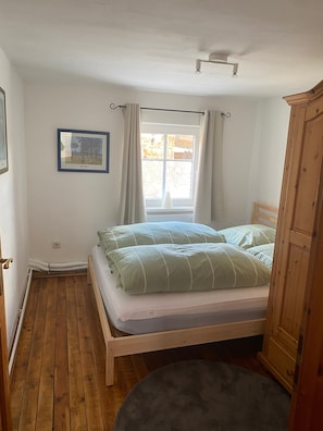 A bedroom with a wooden floor, a bed with green bedding, a window with curtains, a framed picture on the wall, and a wooden wardrobe. - Holiday Home 'Günther' with Shared Terrace, Private Garden and Wi-Fi (Hohen Pritz)