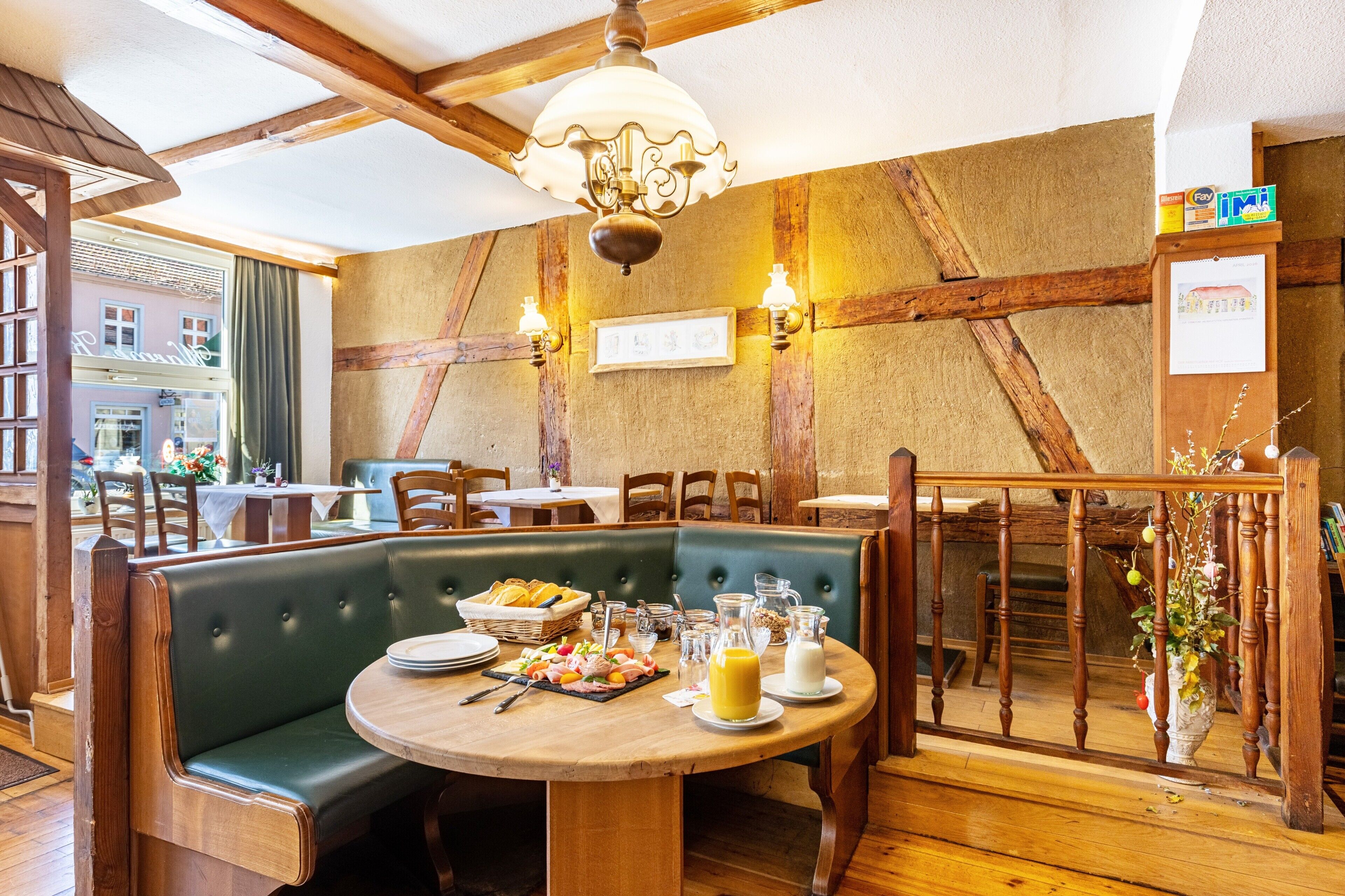 A restaurant with wooden beams, green cushioned seating, a round wooden table with breakfast items, and a hanging light fixture.