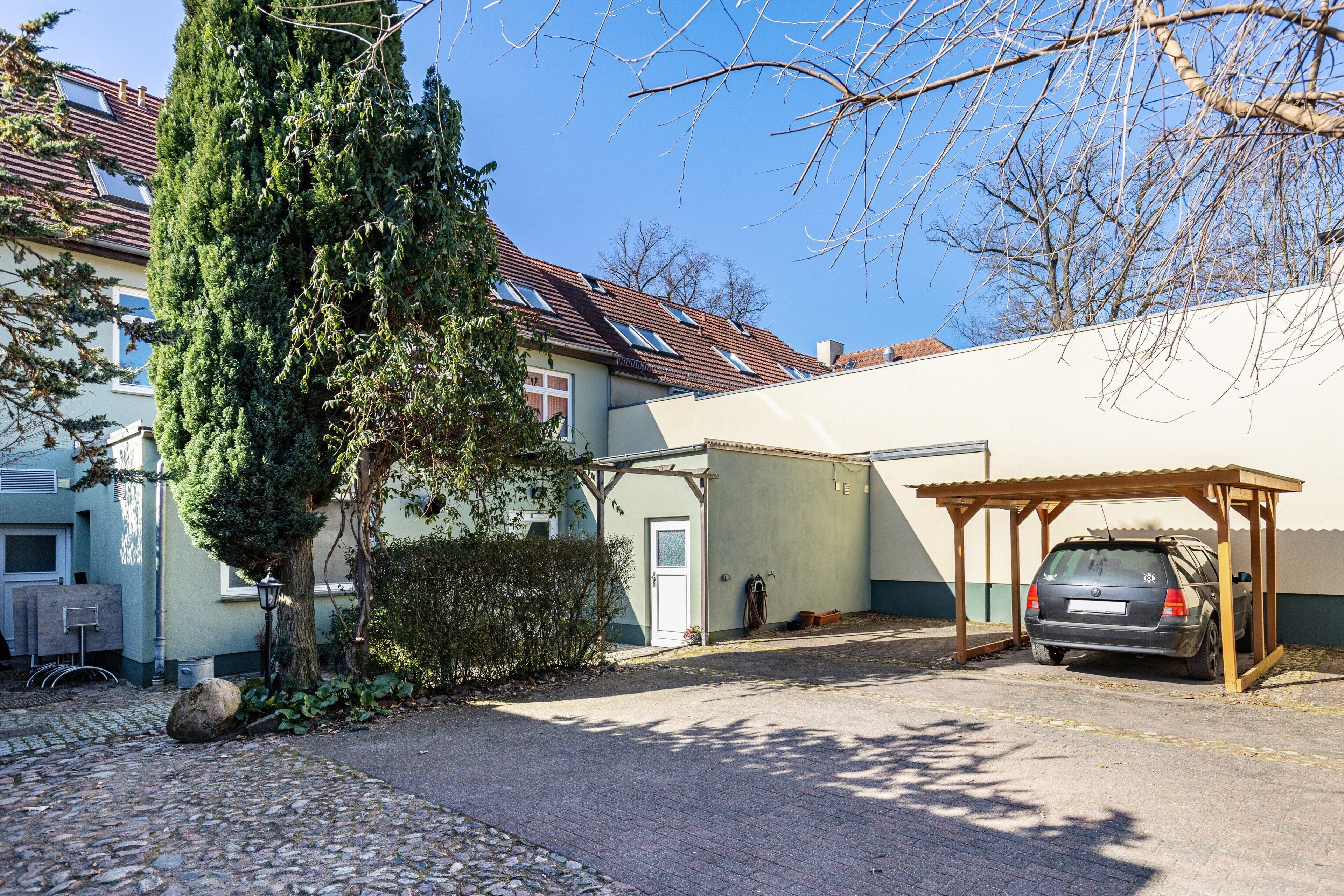 A residential area with a parked car, a covered carport, a stone-paved driveway, and a tall coniferous tree.