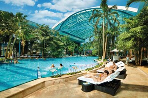 Ein Swimmingpool mit Liegestühlen, Palmen und einem großzügigen Gebäude mit Glasdach.
