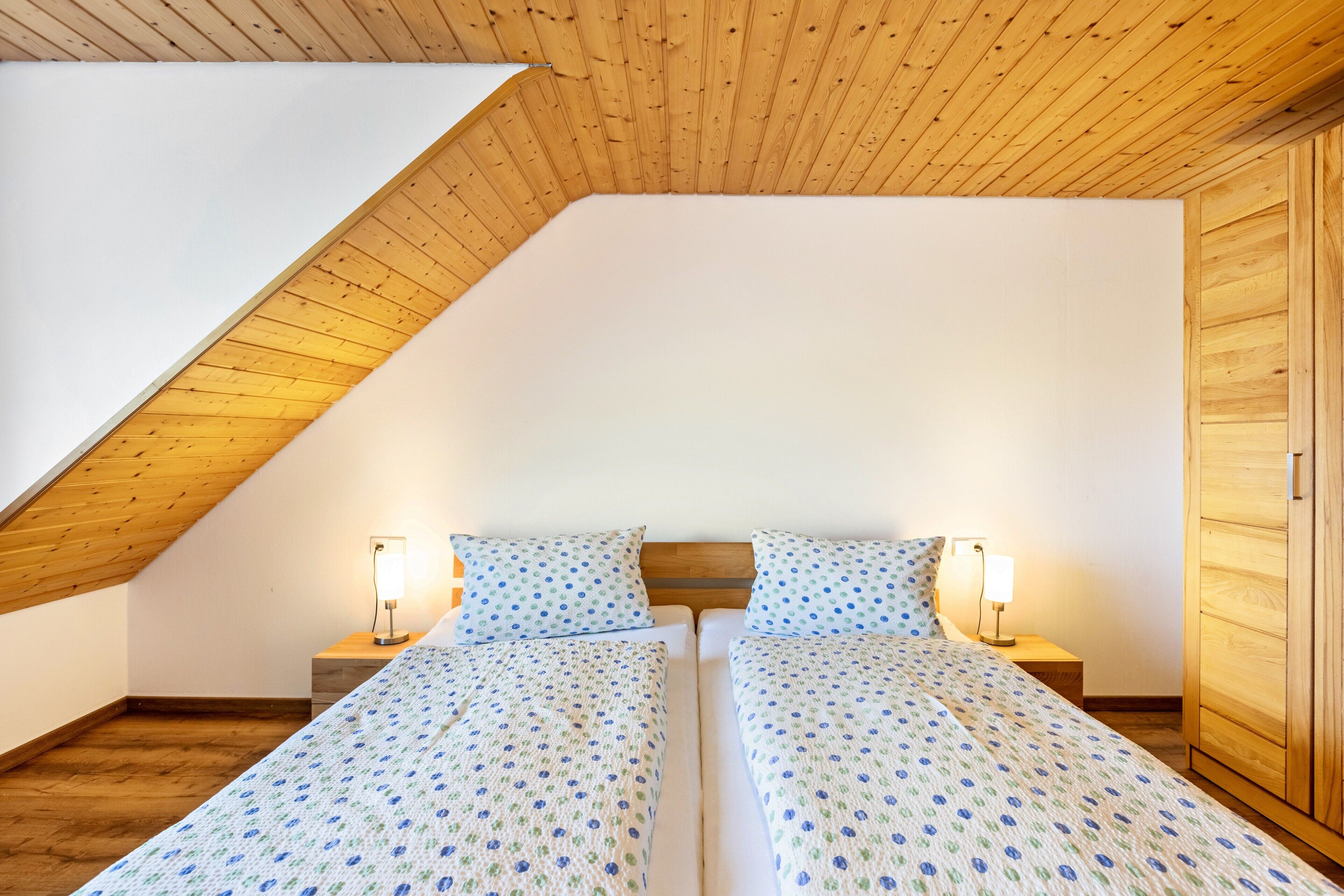 A bedroom with a sloped ceiling, two beds with matching bedding, bedside lamps, and a wooden wardrobe.