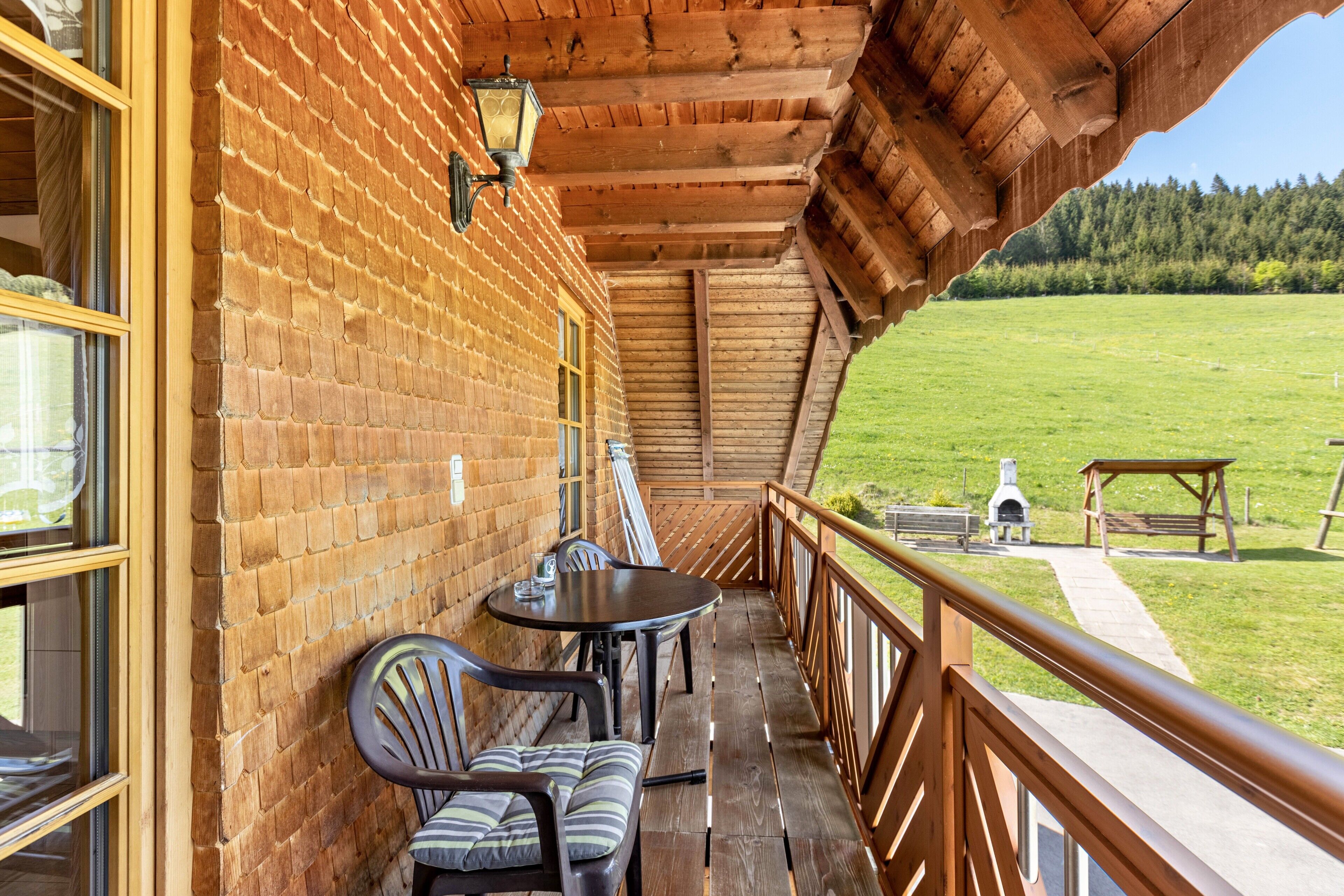 Ein Holzbalkon mit Ziegelwand, Tisch, Stühlen und Blick auf ein Wiesenfeld.
