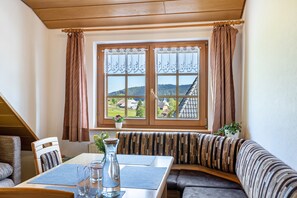Une pièce comprenant une table à manger, un banc, une fenêtre avec des rideaux et une vue sur des maisons et des collines.