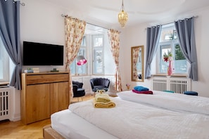 A bedroom with a bed, a TV, a wooden wardrobe, a chair, a desk, and a window with curtains.