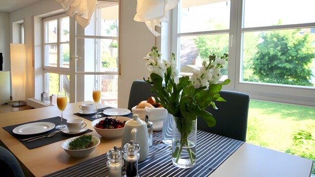 Ein Esstisch ist gedeckt mit Tellern, Tassen, einer Blumenvase und einer Obstschale.