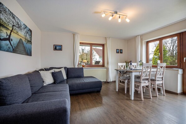 Un salon avec un canapé gris foncé, une table à manger avec des chaises, une fenêtre avec des rideaux et un tableau accroché au mur.