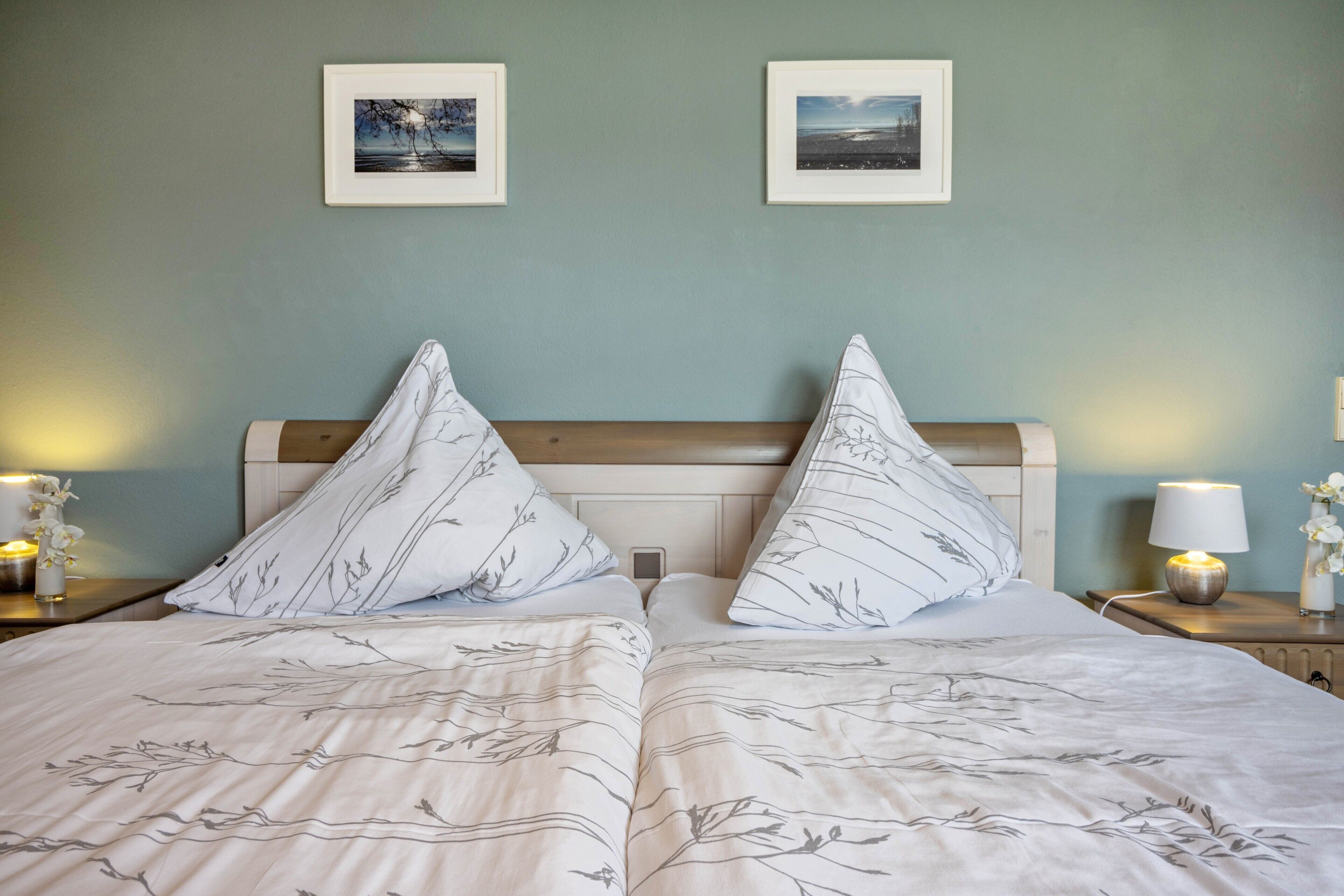 A double bed with a leaf-patterned duvet, two bedside tables with lamps, and two framed pictures on the wall.