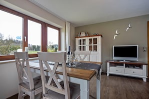 A dining area with wooden chairs and a table, a cabinet with glass doors, a flat-screen TV, and a view of the outdoors.