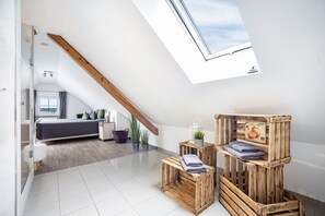 A modern loft bedroom with a skylight, a bed, a small seating area, and a wooden crate storage unit.