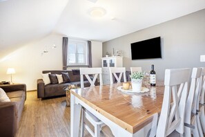 A modern living room with a wooden dining table, white chairs, a sofa, a TV mounted on the wall, and a window with curtains.