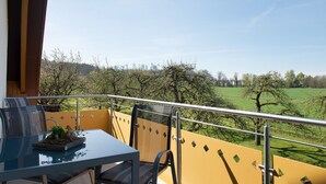 Un balcon avec une table et des chaises, donnant sur un paysage verdoyant avec des arbres et un ciel dégagé.