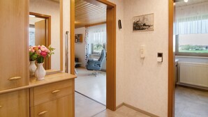 Une chambre d’hôtel avec une commode en bois, un miroir, un vase avec des fleurs, une chaise, un radiateur et une fenêtre donnant sur un beau paysage.