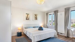 Ein Schlafzimmer mit einem Bett, zwei Nachttischlampen, einem Nachttisch, einem Fenster mit Vorhängen und zwei an der Wand befestigten Kunstwerken.