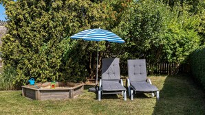 Een tuin met twee loungestoelen, een houten zandbak en een blauw gestreepte parasol.