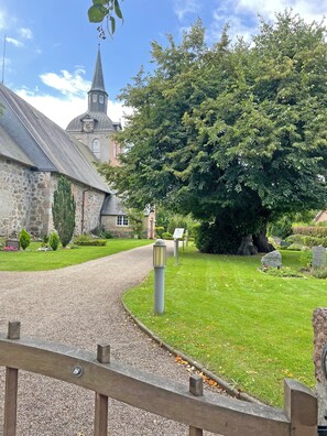 A church with a clock tower, a stone building, and a large tree in a well-maintained garden. - Apartment 'Ferienwohnung Ankerleicht' with Shared Terrace, Shared Garden and Wi-Fi (Steinbergkirche)