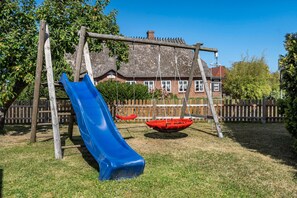 A playground with a blue slide and swings, surrounded by a wooden fence and a house with a thatched roof.