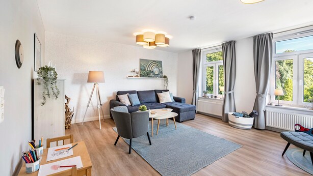 Ein modernes Wohnzimmer mit einer Couch, einem kleinen Tisch und einer Lampe. Ein Fenster mit Vorhängen bietet Blick auf Grünflächen.