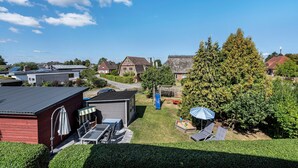 Ein Wohngebiet mit blauem Himmel, Häusern, einem großen Baum und einer Terrasse mit Gartenmöbeln.