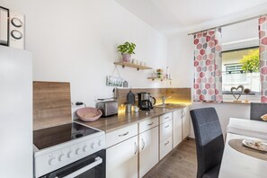 Una cucina moderna con piano cottura, tostapane e macchina da caffè, scaffali in legno con utensili da cucina, e un'area pranzo con una sedia e un tavolo.
