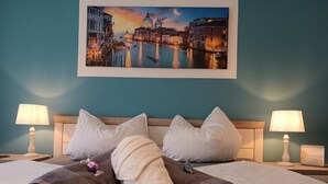 Habitación de hotel con una cama, dos almohadas blancas, dos toallas enrolladas, una mesita de noche con una lámpara y un cuadro de un paisaje urbano.