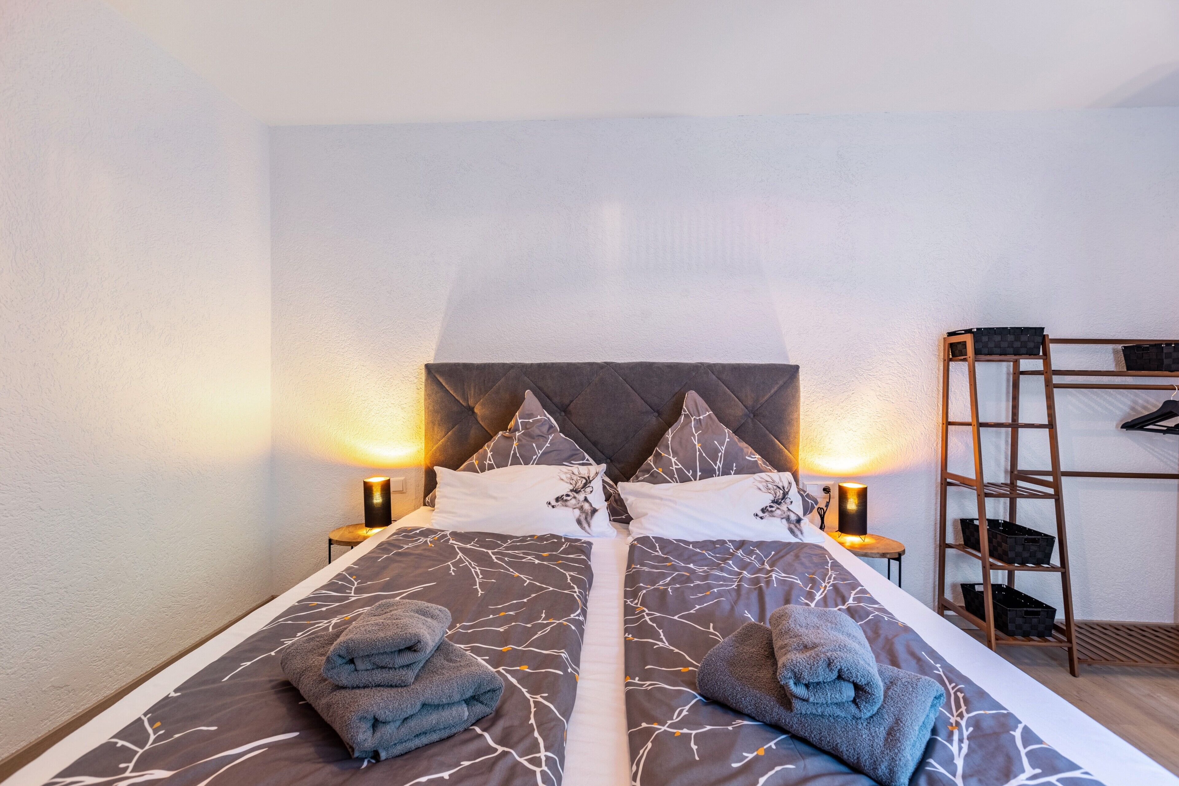 Een tweepersoonsbed met een bedsprei met patroon, twee kussens, twee nachtlampjes en een houten ladder met manden.