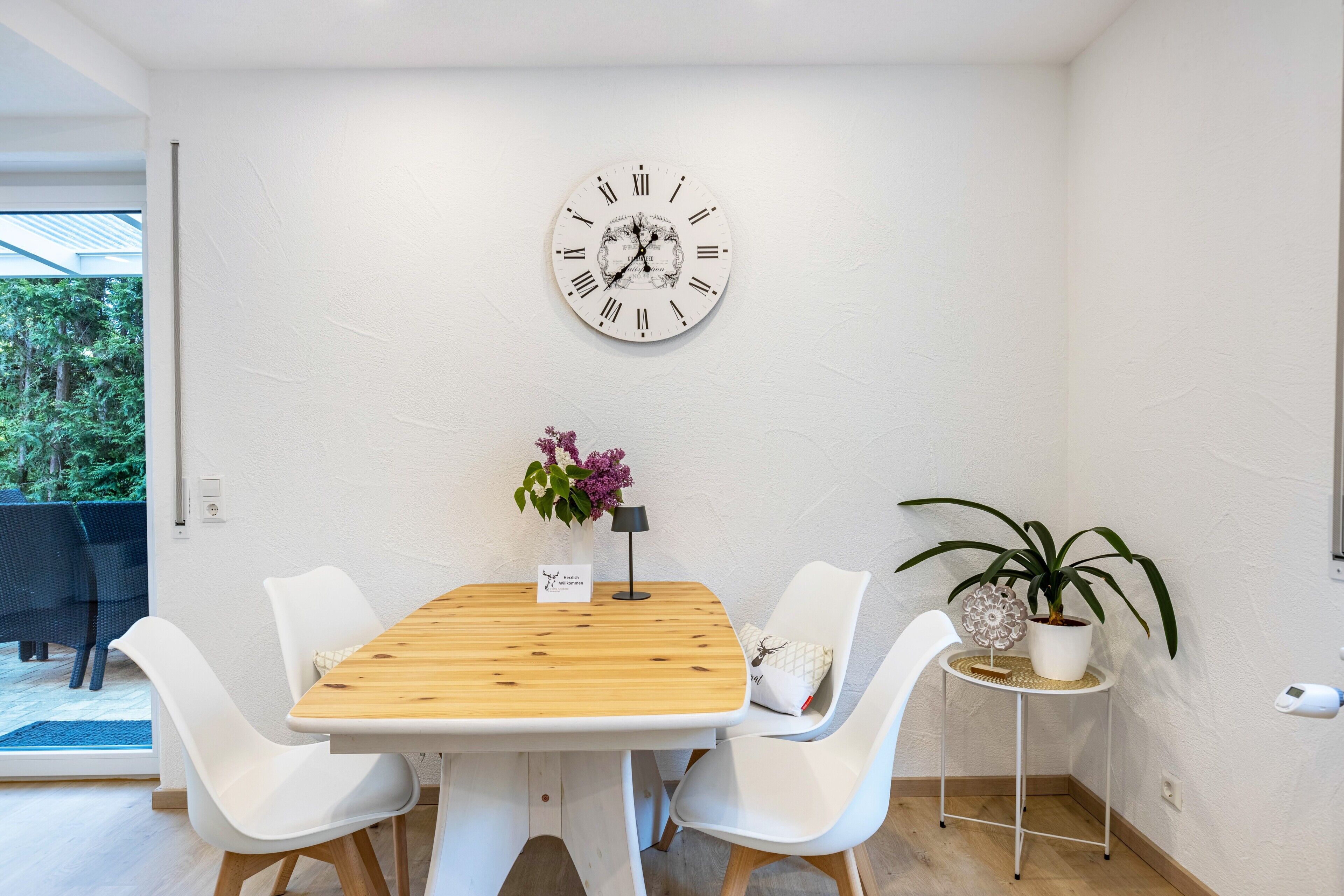 A dining area with a wooden table, white chairs, a wall clock, a small table with a potted plant, and a view of the outdoors.
