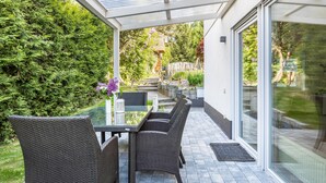 A covered patio with wicker furniture, a glass table, and a view of a garden and house. - Apartment 'Reinbold' with Private Terrace, Private Garden and Wi-Fi (Niedereschach)