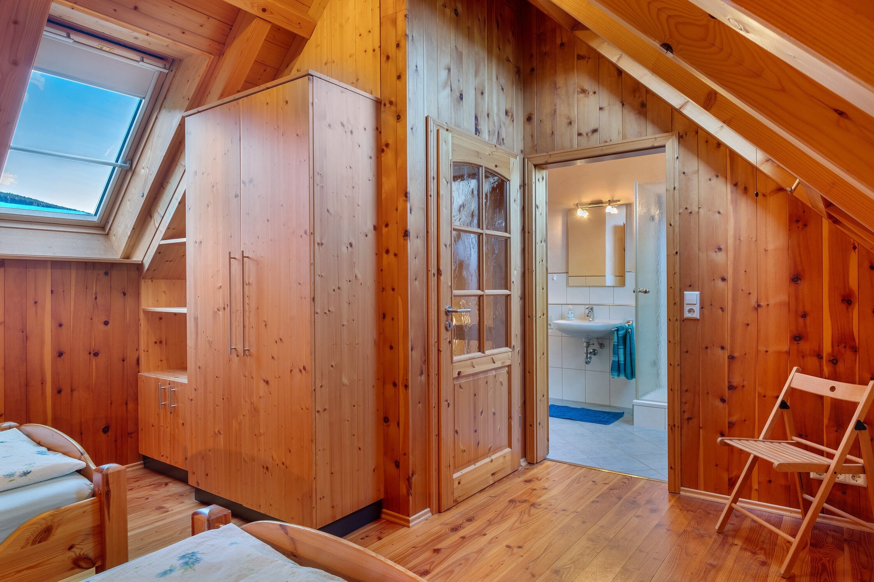 Une pièce en bois avec une lucarne, une armoire, une porte donnant sur une salle de bain et une chaise en bois.
